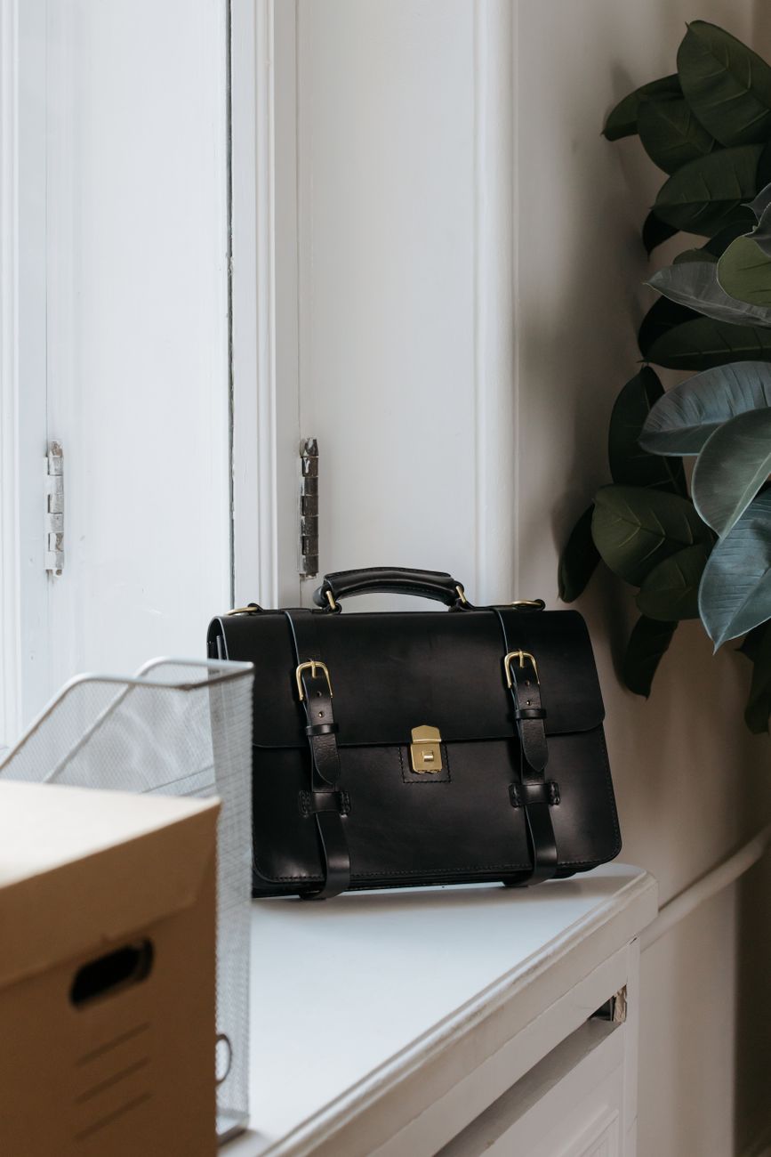 A black leather briefcase closed on a minimalist office desk — the specimen view of a leather laptop bag filed by hide, hardware, and years of use.