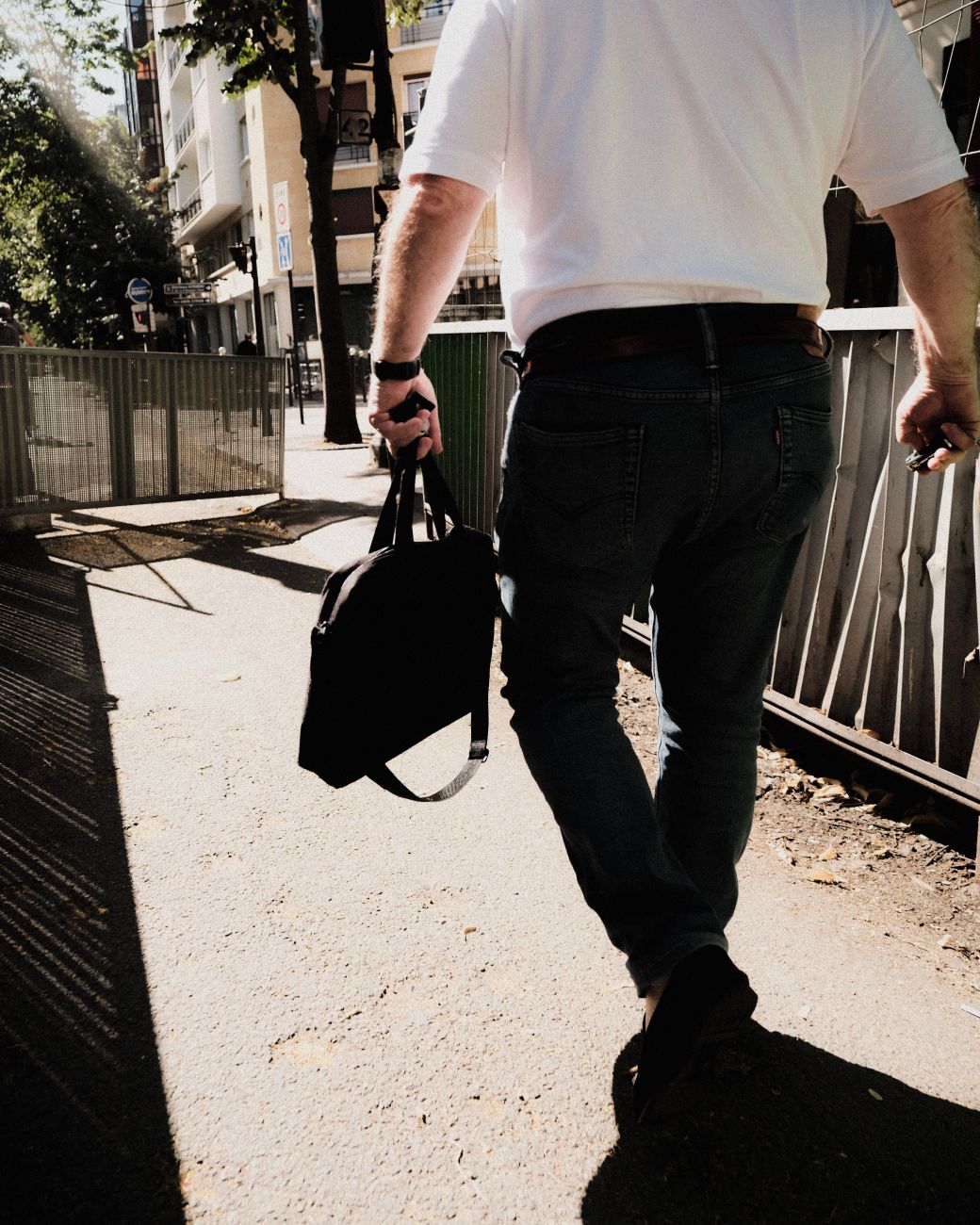 A man walking a sunlit city street with a leather laptop bag over one shoulder — the daily commute that reveals which strap anchor survives seven years.