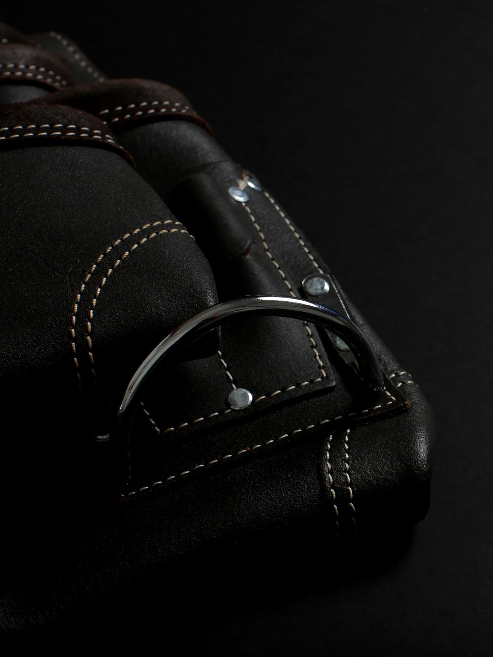 A dark leather laptop bag in raking light with visible saddle-stitching and a solid metal buckle — the maker's mark you can read off construction detail.